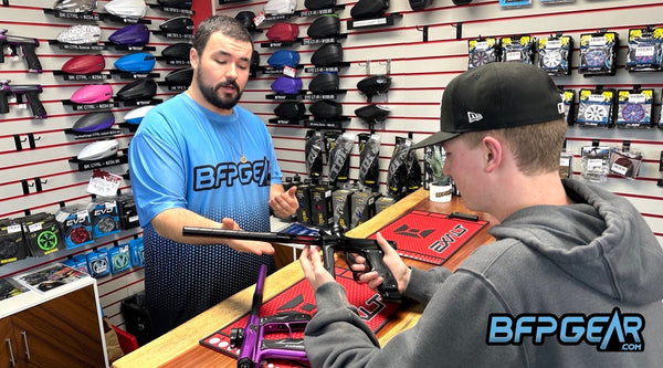 Dufort is pictured here helping a customer pick out a paintball marker. The markers of voice are the Etha 3M, Empire Axe 2.0, and the Shocker AMP.