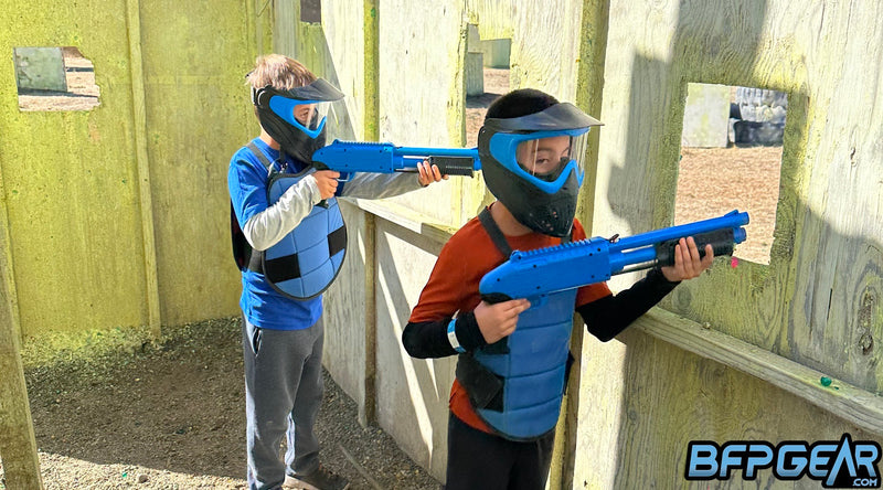 Two players using the JT Splatmaster paintball markers, taking cover inside of a structure. Each player is wearing the proper safety equipment, which are goggles and chest protectors.